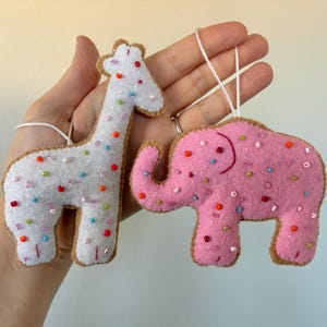 May include: Two felt animal ornaments featuring a white giraffe and a pink elephant. Both figures are decorated with colorful beads and glitter, and each includes a white string loop for hanging. The edges are stitched with tan thread.