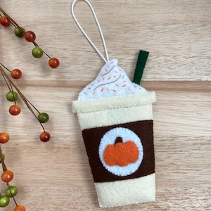 May include: A felt ornament shaped like a coffee cup with whipped cream and sprinkles. The cup has a brown sleeve with a pumpkin design. The ornament has a white string for hanging.