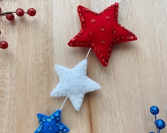 May include: A red, white, and blue star garland with a red star at the top, a white star in the middle, and a blue star at the bottom. The stars are made of felt and have sequins around the edges.