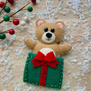 Peut inclure: Une décoration de Noël en feutre représentant un ours en peluche marron qui sort d'une boîte cadeau verte avec un nœud rouge. La boîte cadeau est décorée de paillettes vertes.
