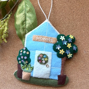 May include: A handmade felt ornament shaped like a blue house with the word "FLOWERS" on the roof. The house has a white door with a floral wreath, and is flanked by felt trees and potted plants. A white string is attached for hanging.