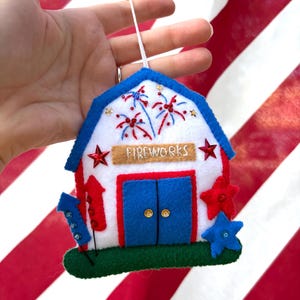 Può includere: Un ornamento in feltro a forma di fienile con facciata bianca, bordi blu, ricami di fuochi d'artificio rossi e blu e un cartello color sabbia con la scritta FIREWORKS. La struttura presenta porte blu con paillettes dorate e stelle decorative.