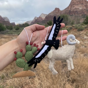 Op de afbeelding: Handgemaakte vilten ornamenten met een cactus, een zwart-witte vogel en een ram. De cactus is groen met rode accenten, de vogel heeft een witte streep en de ram is beige. Elk ornament heeft een wit touwtje om op te hangen.