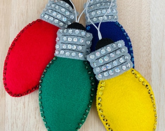 Puede incluir: Cuatro adornos navideños de fieltro con forma de bombilla. Las bombillas son rojas, verdes, azules y amarillas. Cada bombilla tiene una parte superior plateada y gris con pedrería.