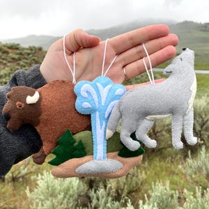 Può includere: Ornamenti in feltro fatti a mano raffiguranti un bisonte marrone, un geyser blu e un lupo grigio. Ogni ornamento ha un cordino bianco per appenderlo. Il bisonte e il lupo sono in stile naturalistico, il geyser è stilizzato.