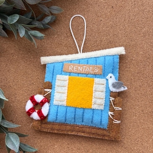 May include: A felt hanging ornament shaped like a blue beach shack with a yellow window, a white and red life preserver, and a small white seagull. The sign above the window reads RENTALS in white thread. It includes a white string loop.