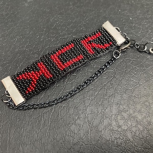 May include: Black beaded bracelet with the word "MCR" in red. The bracelet has silver-colored metal end pieces and a black chain with a fleur-de-lis charm. The bracelet is on a dark gray surface.