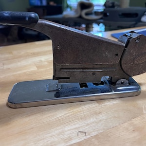 May include: Vintage stapler with a black handle and a silver base. The stapler has a worn, rusty appearance, suggesting age and use. The design is classic, with a curved body and a circular plate on the side. It sits on a light-colored wooden surface.