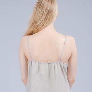 May include: A woman wearing a light beige linen dress with thin straps. The dress is loose fitting and has a gathered neckline.