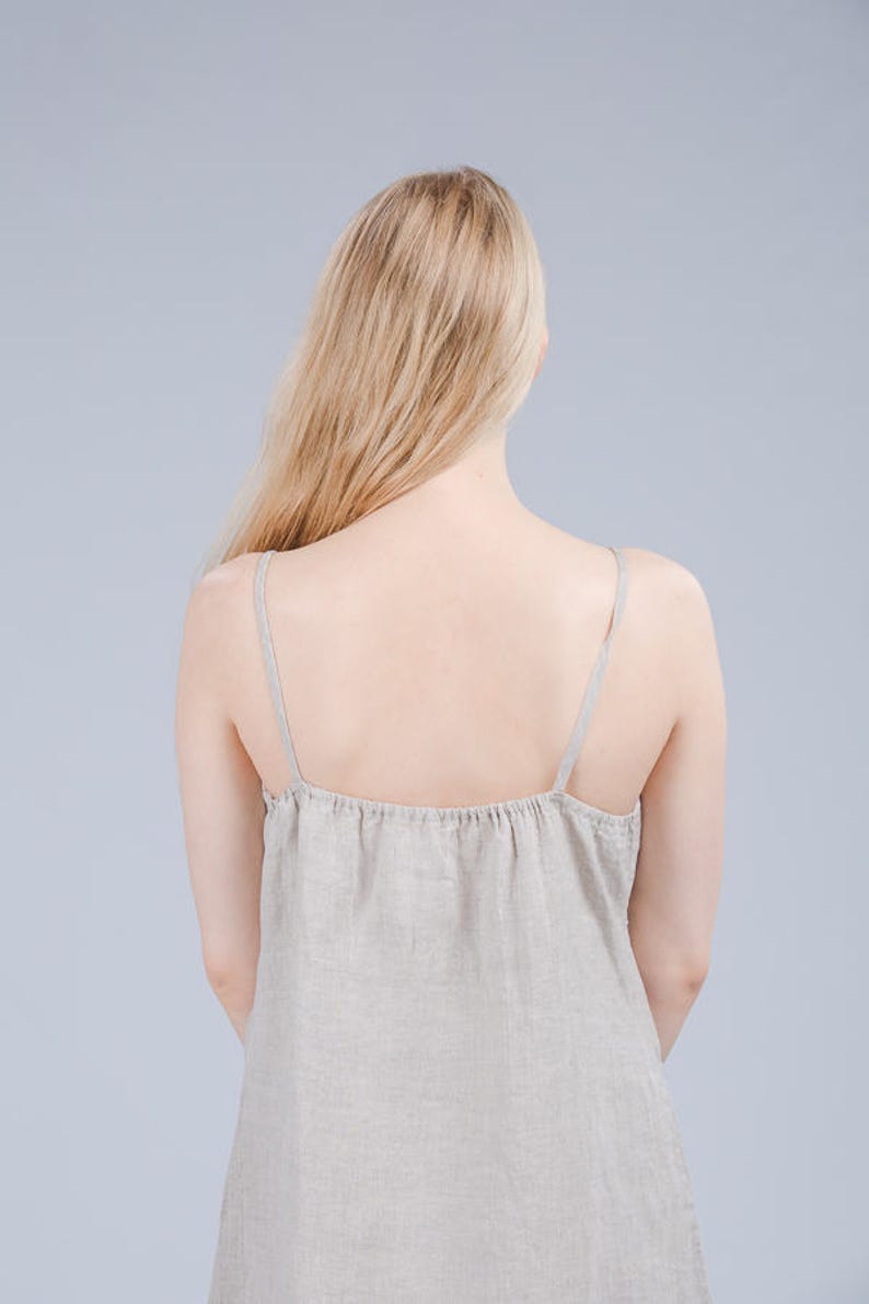 May include: A woman wearing a light beige linen dress with thin straps. The dress is loose fitting and has a gathered neckline.