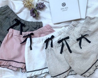 May include: Four pairs of linen shorts with black ribbon ties and lace trim. The shorts are in gray, pink, white, and striped. The shorts are arranged on a white fabric background. The text "OMALINEN natural" is visible on a white box.