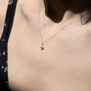 May include: A delicate gold chain necklace featuring a single, small, round white pearl pendant. The necklace rests against the collarbone, showcasing a thin, polished gold metal chain that holds the pearl securely in a simple gold setting.