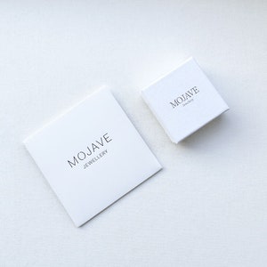 May include: Two square white gift boxes of different sizes, each featuring the black text MOJAVE JEWELLERY printed on the lid. The boxes have a textured finish and are positioned at an angle, showcasing a clean and simple design for packaging.