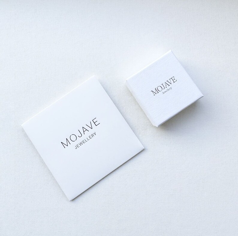 May include: Two square white gift boxes of different sizes, each featuring the black text MOJAVE JEWELLERY printed on the lid. The boxes have a textured finish and are positioned at an angle, showcasing a clean and simple design for packaging.