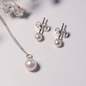 May include: A jewelry set consisting of a silver chain necklace with a single round white pearl pendant and a pair of matching silver stud earrings, each featuring a small round white pearl. The metal components are polished silver.
