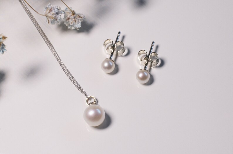 May include: A jewelry set consisting of a silver chain necklace with a single round white pearl pendant and a pair of matching silver stud earrings, each featuring a small round white pearl. The metal components are polished silver.