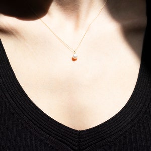 May include: A delicate gold chain necklace featuring a single, small, round white pearl pendant. The necklace rests against the skin, with the thin metallic chain extending upwards from the pearl, which is centered at the base of the neck.