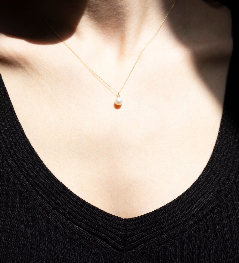 May include: A delicate gold chain necklace featuring a single, small, round white pearl pendant. The necklace rests against the skin, with the thin metallic chain extending upwards from the pearl, which is centered at the base of the neck.