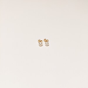 May include: A pair of stud earrings featuring round white pearls mounted on gold-toned metal posts. The earrings are shown from the front, displaying the smooth, spherical surface of the pearls and the metallic backing of the earring posts.