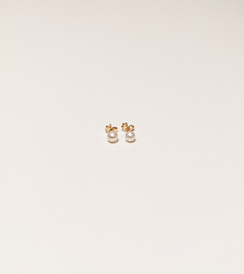 May include: A pair of stud earrings featuring round white pearls mounted on gold-toned metal posts. The earrings are shown from the front, displaying the smooth, spherical surface of the pearls and the metallic backing of the earring posts.