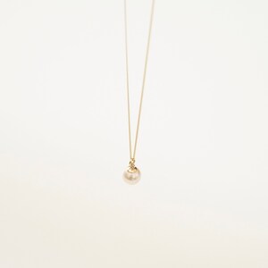 May include: A delicate gold chain necklace featuring a single, round, cream-colored pearl pendant. The pearl is suspended from a small gold-toned metal cap that connects directly to the thin, minimalist gold chain, creating a simple, elegant look.