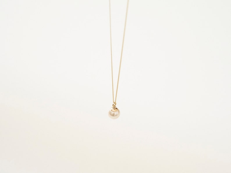 May include: A delicate gold chain necklace featuring a single, round, cream-colored pearl pendant. The pearl is suspended from a small gold-toned metal cap that connects directly to the thin, minimalist gold chain, creating a simple, elegant look.