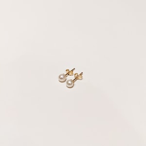 May include: A pair of stud earrings featuring small, round, white pearls set on gold posts. Each earring includes a gold butterfly-style backing, showcasing a classic design with a polished metallic finish and smooth, spherical white pearls.