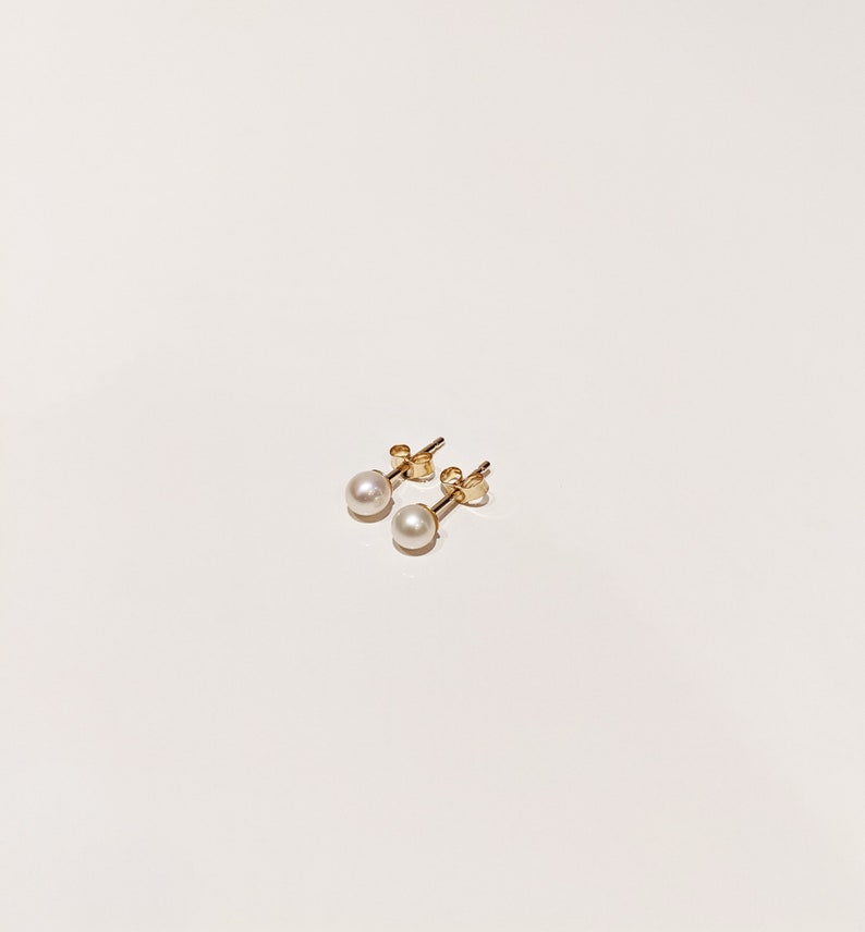 May include: A pair of stud earrings featuring small, round, white pearls set on gold posts. Each earring includes a gold butterfly-style backing, showcasing a classic design with a polished metallic finish and smooth, spherical white pearls.