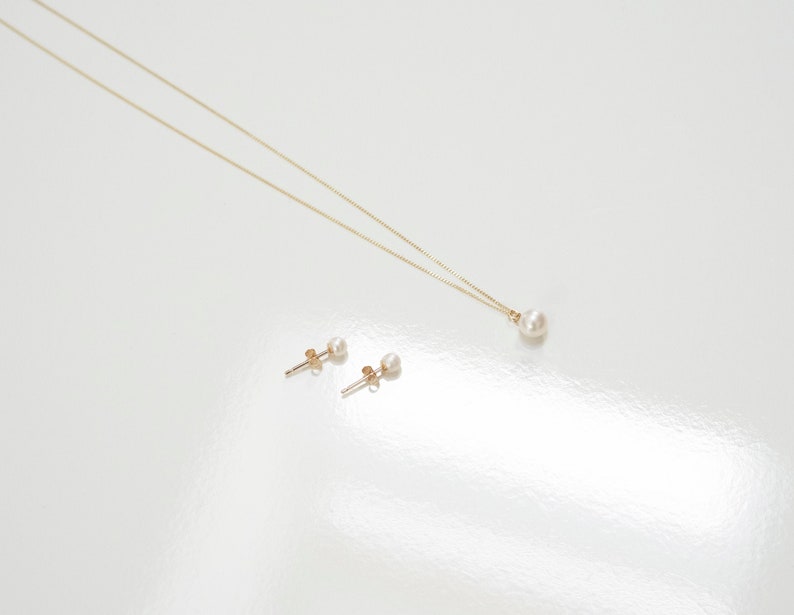 May include: A gold chain necklace featuring a single round white pearl pendant and a pair of matching gold stud earrings, each set with a small round white pearl. The jewelry set is displayed against a plain, light-colored surface.