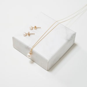 May include: A gold chain necklace featuring a single round white pearl pendant and a pair of matching pearl stud earrings with gold posts. The jewelry set is displayed together, showcasing the delicate gold metal links and the smooth, lustrous finish of the pearls.