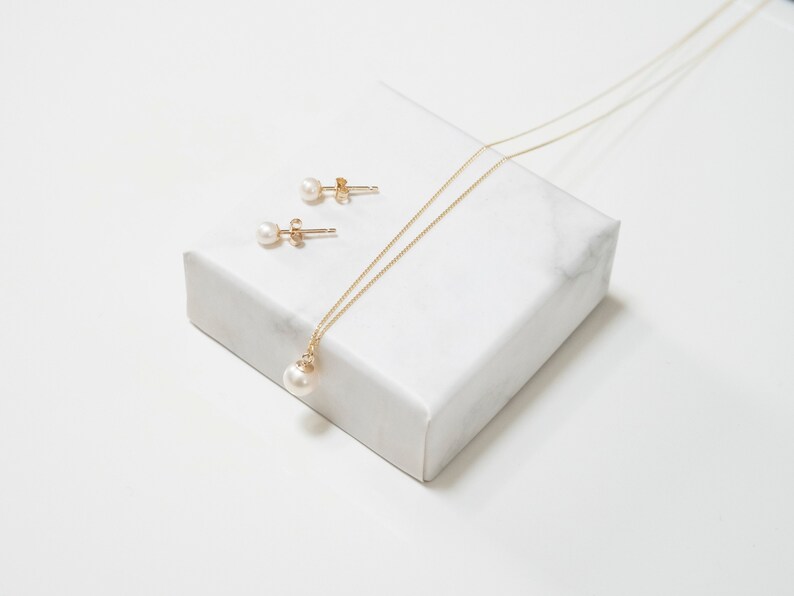 May include: A gold chain necklace featuring a single round white pearl pendant and a pair of matching pearl stud earrings with gold posts. The jewelry set is displayed together, showcasing the delicate gold metal links and the smooth, lustrous finish of the pearls.