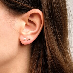 May include: A pair of gold-toned earrings consisting of one small round white pearl stud and one thin, horizontal gold-toned bar stud. Both earrings are worn in the earlobe, featuring a simple, polished design with a metallic finish and a smooth pearl.