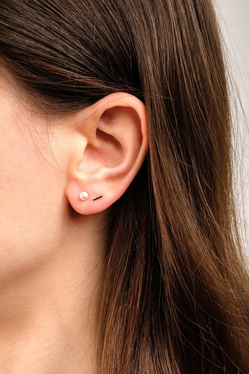 May include: A pair of gold-toned earrings consisting of one small round white pearl stud and one thin, horizontal gold-toned bar stud. Both earrings are worn in the earlobe, featuring a simple, polished design with a metallic finish and a smooth pearl.
