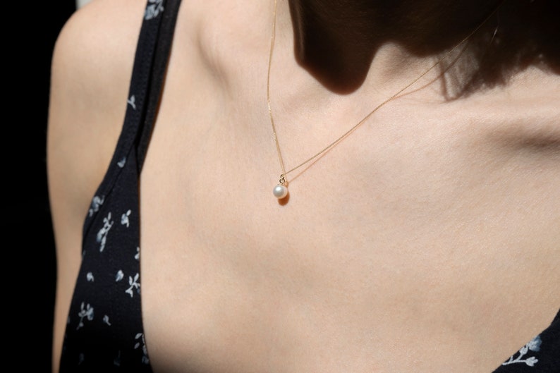 May include: A delicate gold chain necklace featuring a single, small, round white pearl pendant. The necklace rests against the collarbone, showcasing a thin, polished gold metal chain that holds the pearl securely in a simple gold setting.