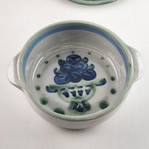 Puede incluir: Un cuenco de cerámica con interior gris, detalles azules y verdes y dos asas. El cuenco presenta un diseño floral azul en el centro y lunares verdes alrededor del borde. El borde tiene una franja azul y verde.