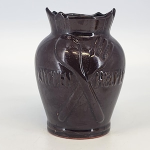 May include: A dark brown ceramic kitchen utensil holder. The vase-shaped holder features embossed crossed spoon and fork designs. The words "Kitchen Help" are embossed on the front. The top edge has a scalloped design.