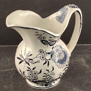 May include: White ceramic pitcher with a curved handle and a scalloped rim. The pitcher is decorated with blue floral patterns and foliage. The base and rim are outlined in blue. A decorative piece for the kitchen or dining room.
