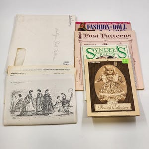 May include: A collection of vintage doll patterns and related ephemera. Includes a booklet titled "Fashion Doll," a "Portrait Collection" booklet featuring a doll, and various pattern instructions. The items are arranged on a white surface.