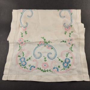 Puede incluir: Un paño de lino blanco con diseños florales bordados. El bordado presenta flores rosas y azules, hojas verdes y detalles azules en forma de volutas. El paño tiene un borde dobladillado.