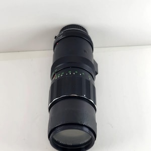 May include: A black zoom lens with a leather-like grip and various measurement markings in green and white. The lens has a silver ring near the front and a clear glass element. The lens is designed for photography.