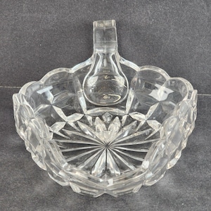 Może przedstawiać: Przezroczysta szklana miska z uchwytem, ozdobiona szlifowanym wzorem. Miska ma kwadratowy kształt z zaokrąglonymi rogami i falistym brzegiem.