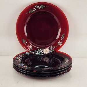 May include: A stack of four burgundy-colored glass plates with a floral design. The plates have a dark red hue and are decorated with white, pink, and blue flowers and green leaves around the rim. The plates are stacked on top of each other.