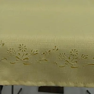 Könnte beinhalten: Hellgelber Stoff mit einem floralen Ausschnittmuster am unteren Rand. Der Stoff hat eine subtile Webstruktur. Das Ausschnittmuster zeigt stilisierte Blumen und Blätter und verleiht dem Textil eine zarte Note.