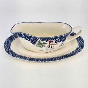 May include: Cream-colored gravy boat and saucer set with a blue dotted border. The gravy boat features a winter scene with a snowman and a Christmas tree. The set is made of ceramic.
