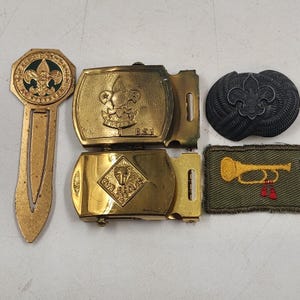 May include: A collection of vintage Boy Scout and Cub Scout memorabilia. Includes a gold-colored bookmark, two belt buckles, a black emblem, and a patch with a gold bugle design on a green background.