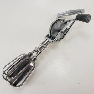 May include: Vintage hand-crank egg beater made of stainless steel and chrome. The beater features a gear mechanism, a handle, and two wire beaters. The word "STAINLESS" is visible on the beater. A classic kitchen tool.
