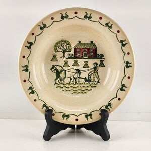 May include: A cream-colored ceramic soup bowl with a green and red farmhouse scene. The scene features a red house, a tree, two horses pulling a plow, and a farmer. The bowl has a green and red scalloped edge.