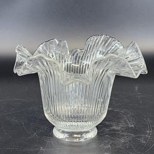 Puede incluir: Jarrón de cristal transparente con borde ondulado y diseño de estrías verticales. El jarrón tiene una base redonda y una parte superior acampanada y ondulada. El cristal transparente permite el paso de la luz, y la forma recuerda a una flor.