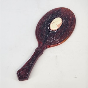 May include: A vintage hand mirror with a dark brown, ornate frame. The oval mirror features a cameo in the center. The handle is also decorated with floral patterns. A classic vanity accessory.