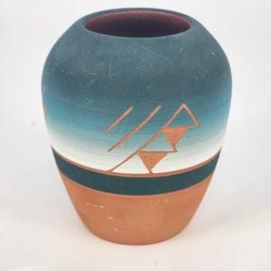 May include: A small, round ceramic vase with a gradient of teal and white, transitioning to a terracotta base. The vase features a geometric design in a lighter terracotta colour. The interior is a dark reddish-brown.